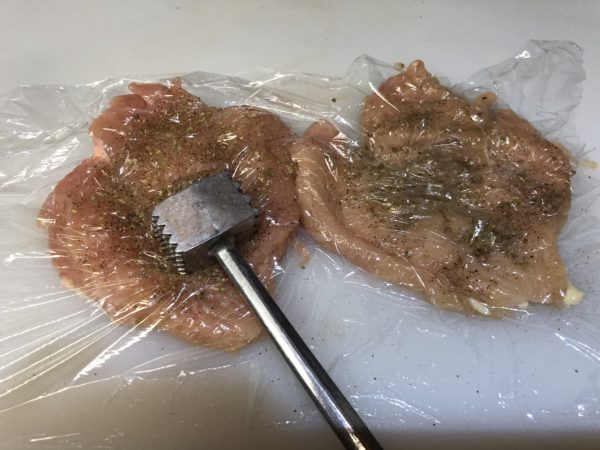 Flattening seasoned chicken breasts with plastic wrap and meat mallet. Use the flat side of mallet by pounding chicken until even thickness.